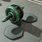 A green and black ab roller exercise wheel with a handle, shown on a speckled dark gray gym floor alongside two textured floor mats. The brand name 'STUDIO' is visible on the roller.