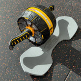 A black and yellow ab roller wheel exercise device resting on a gray bone-shaped foam pad over a speckled black gym floor.