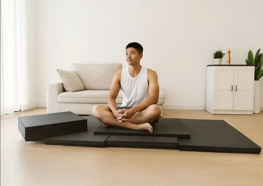 Man sitting on stacked black gym mats in a bright living room, preparing for a home workout or stretching session