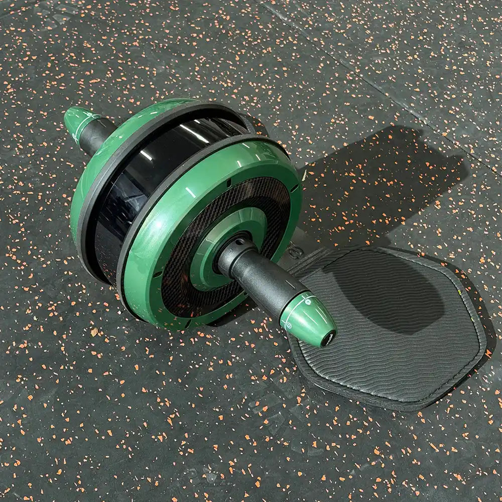 A green and black ab roller wheel exercise device with a black textured mat, shown on a dark speckled gym floor.