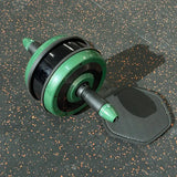 A green and black ab roller wheel exercise device with a black textured mat, shown on a dark speckled gym floor.