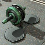A green and black ab roller exercise wheel with a handle, shown on a speckled dark gray gym floor alongside two textured floor mats. The brand name 'STUDIO' is visible on the roller.