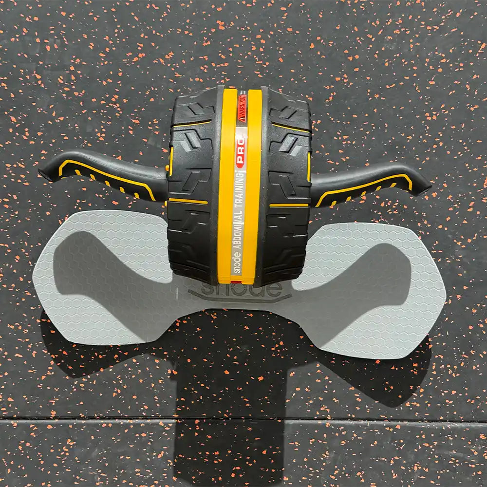 An abdominal exercise wheel with black handles and a black central body featuring a bold yellow stripe, resting on a gray wheeled support pad over a black rubberized gym floor.