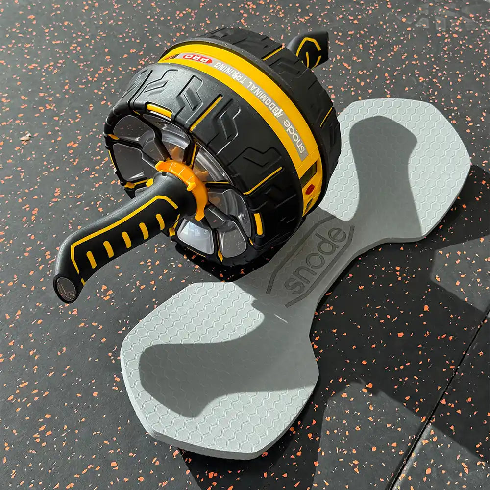 A black and yellow ab roller wheel exercise device resting on a gray bone-shaped foam pad over a speckled black gym floor.