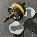 A black and yellow ab roller wheel exercise device resting on a gray bone-shaped foam pad over a speckled black gym floor.