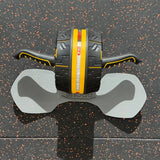 An abdominal exercise wheel with black handles and a black central body featuring a bold yellow stripe, resting on a gray wheeled support pad over a black rubberized gym floor.