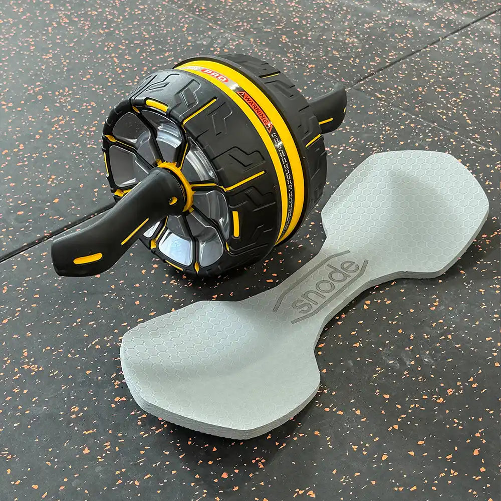 An ab roller wheel exercise device in black and yellow, shown alongside a light gray foam roller on a dark speckled gym floor. The ab roller features a textured tread pattern and a black handle, while the foam roller has the brand name 'snode' printed on it.