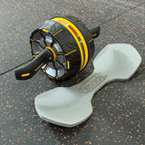 An ab roller wheel exercise device in black and yellow, shown alongside a light gray foam roller on a dark speckled gym floor. The ab roller features a textured tread pattern and a black handle, while the foam roller has the brand name 'snode' printed on it.