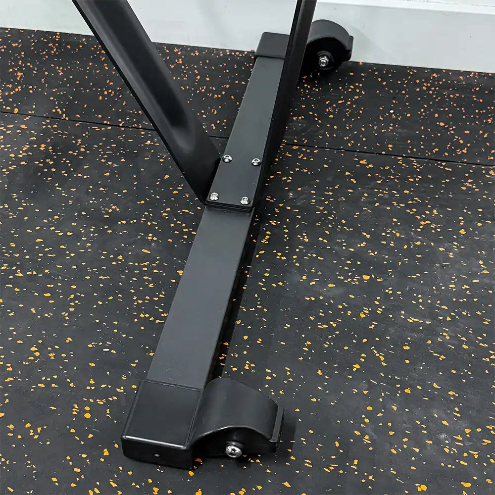 Close-up of a black metal equipment frame with caster wheels on a black rubber gym floor with orange and yellow speckles.