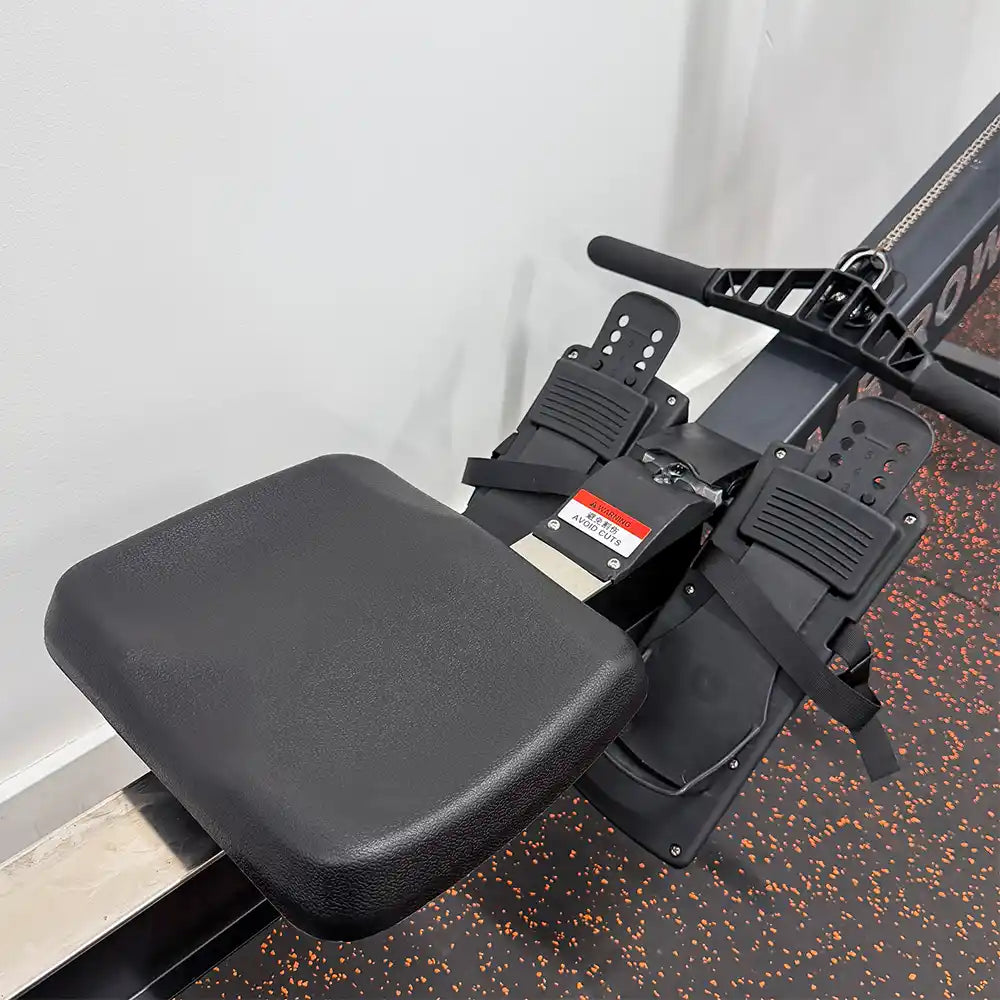 Close-up of a rowing machine seat and foot pedals in black, shown on a gym floor with speckled dark rubber and a light wall in the background.